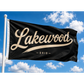 A black flag with 'Lakewood - Ohio' printed in beige script and serif lettering, flying on a white flagpole against a bright blue sky with white clouds.