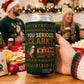 A hand holds a green can cooler featuring a Christmas-themed graphic that reads 'YOU SERIOUS, CLARK?' with a camper illustration. The background shows a festive holiday gathering with people in sweaters and Santa hats, holding matching can coolers.
